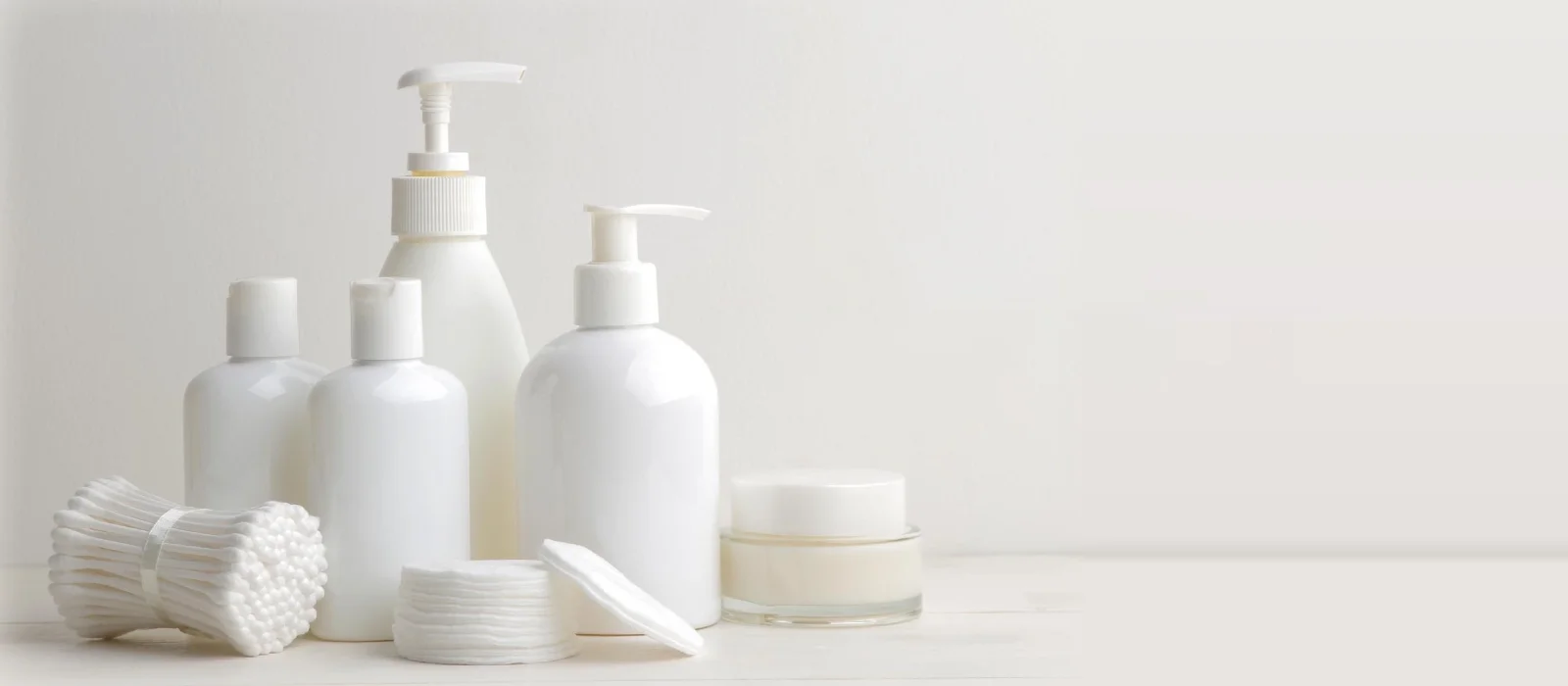 A collection of skincare products in white bottles with pump dispensers, accompanied by cotton pads and swabs on a light background.